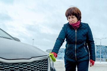Elderly woman wiping front of gray car with microfiber rag on snowy urban lot under cloudy sky, showing responsible driver attitude, winter auto care and active retirement lifestyle themes.