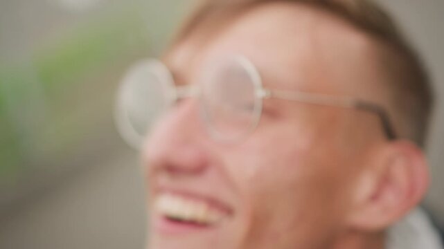 young white man side profile smiling, wet round glasses with raindrop reflections, green blurred park background, wind tussled hair, candid laughter, casual jacket, playful commuter energy, roles