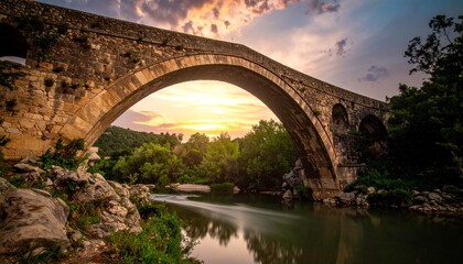 Ancient stone bridge arching over a tranquil river at sunset with lush green trees and dramatic clouds.