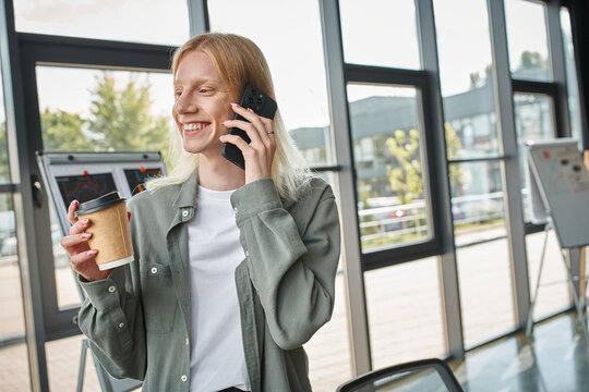 Young non binary person enjoys coffee while chatting on the phone in a bright office space