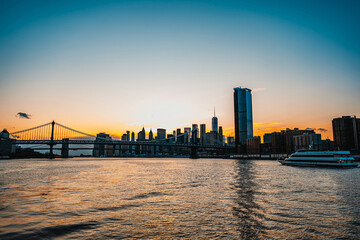 Obraz premium Beautiful sunset over Manhattan skyline and river. The sun sets behind the Manhattan skyline, highlighting bridges and buildings along the river's edge in vibrant colors.