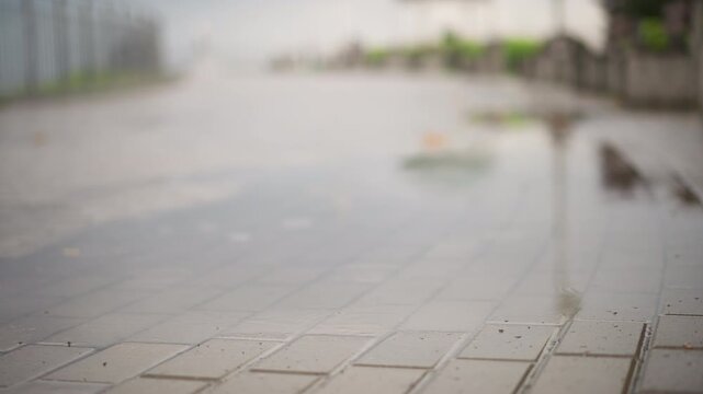wet tiled sidewalk reflective surface shallow, empty urban promenade with puddles and glossy tiles, blurred distant lamppost and fence, overcast sky, muted tones, calm deserted atmosphere, texture