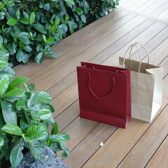 Two shopping bags in red and tan sitting on a wooden deck by green bushes.
