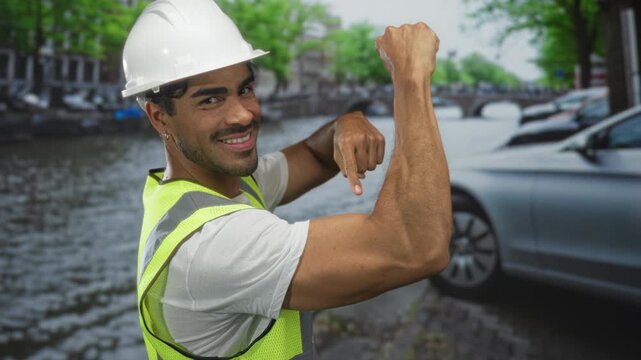 Young hispanic man engineer flexes bicep and points finger beside river street; confidence strength.