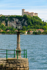 Lamp post on a lakeside pier in Angera, with Lake Maggiore waters in the foreground and a medieval castle on a green cliff under a clear blue sky, northern Italy.
