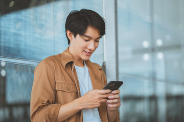 Happy young Asian man in a brown shirt using a smartphone while leaning against a glass wall in a modern building or airport terminal