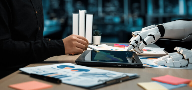 A futuristic workspace showcasing a human hand and a robotic hand collaborating on digital analysis using a tablet, surrounded by productivity tools and documents.Noogenesis - Powered by Adobe
