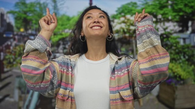Woman pointing fingers upward with visible hands and smiling face on street with green park foliage, wearing patterned jacket and hoop earrings; joy.