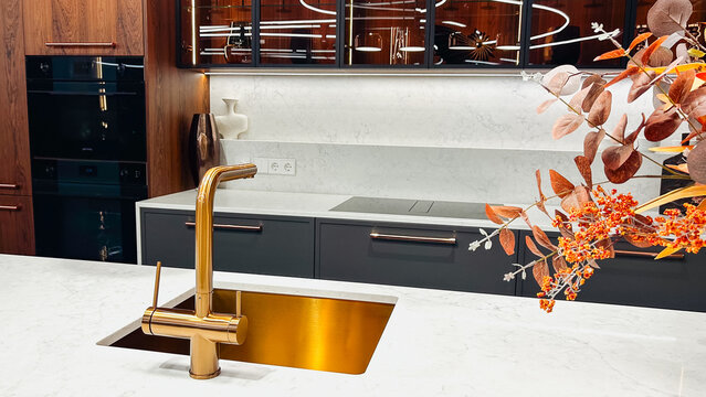Contemporary luxury kitchen with golden faucet, white marble countertop, dark cabinets, elegant autumn foliage.