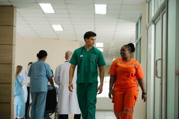two healthcare professionals walking and talking in a hospital corridor. One is wearing green medical scrubs with a stethoscope, and the other is in bright orange scrubs, smiling as they converse.