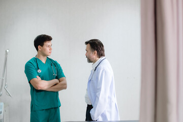 two male doctors having a discussion in a hospital room. One doctor, dressed in green scrubs with a stethoscope, stands with his arms crossed while listening attentively. The other doctor, in a white 