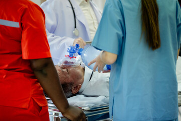 an elderly patient lying on a hospital stretcher receiving emergency medical treatment. Healthcare professionals in medical uniforms are using a manual resuscitator (Ambu bag) to provide assisted vent