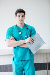 A confident young doctor in green scrubs stands with folded arms, wearing a stethoscope, smiling slightly, representing professionalism and trust in modern healthcare.