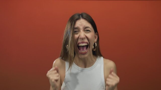 Woman clenches fists in orange studio lighting, eyebrows furrowed, jaws clenched, shoulders raised and lips parted; anxiety.