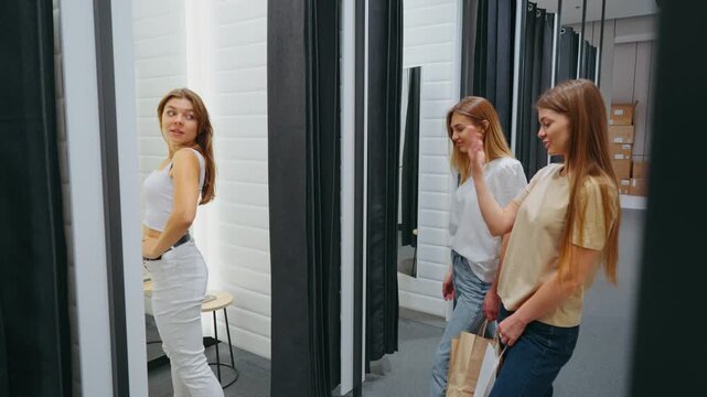 Woman stands outside fitting room showing new outfit to two friends who give feedback, concept of fashion choice and shopping experience