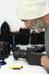 A man in a white beanie and black gloves carefully inspects a DSLR camera with a wide angle lens attached. Precision tools and cleaning supplies are organized on the white table before him.