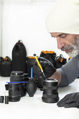 A bearded man in a white beanie and black gloves uses a precision screwdriver to repair a camera lens. Other lenses, a blower, and cleaning tools sit on his white workspace.