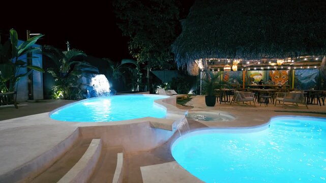 Beautiful swimming pool and jacuzzi illuminated with blue lights at night in a tropical luxury hotel resort with a small waterfall