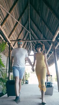 Smiling couple holding hands and walking with their suitcases through a tiki style hotel corridor. Happy tourists checking in for vacation. Vertical