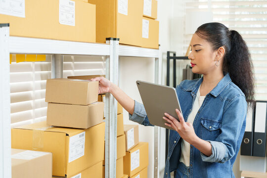 Asian woman entrepreneur checking parcel box labels while holding tablet inside home warehouse, small business online selling, order fulfillment process, shipping preparation, inventory tracking