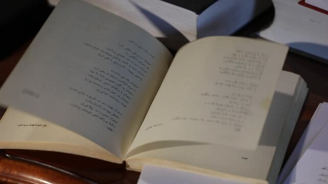 High angle close-up view of an open book containing Persian poetry. The pages with traditional calligraphy text move slightly in the breeze under dim indoor lighting.

