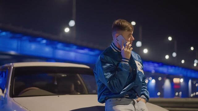 Man on phone under bridge, Individual in bomber jacket communicates beneath glowing bridge lights, Tense discussion takes place beneath bridge illuminated by neon lights and traffic bokeh