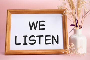 We listen symbol. Concept words We listen on beautiful wooden picture frame. Beautiful pink background with flower vase. Business and We listen concept. Copy space.