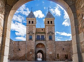 A grand view of the Puerta de Bisagra gate in Toledo, Spain, featuring two stone towers with tiled pyramidal spires and a central archway under a bright cloudy sky