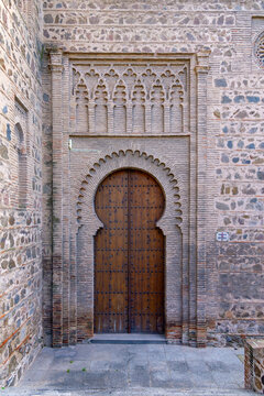 An intricate Mud&eacute;jar brick horseshoe arch frames a studded wooden door at Santiago del Arrabal in Toledo, Spain, set against ancient stone and decorative masonry