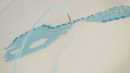 A close-up view of a needle stitching light blue thread onto a fabric, revealing a partially completed embroidery pattern. The intricate detail highlights the craftsmanship involved