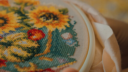 A close-up view of vibrant cross-stitch embroidery featuring sunflowers, with intricate details in red, yellow, and green threads. The fabric is held in a circular embroidery hoop