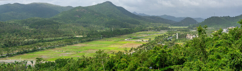 Panoramic high angle view of green agricultural fields in a valley at the foot of tropical mountains under cloudy sky scenic Hainan island rural landscape for ecotourism and agronomy.