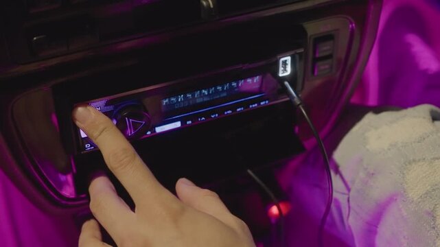 night car stereo finger pressing playlist buttons, usb cable plugged into port, towel draped over passenger seat, magenta neon glow bathing dashboard, tactile navigation through tracks, intimate urban