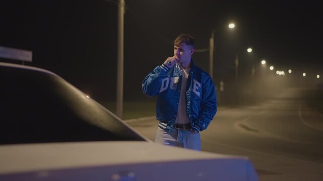 night roadside scene white male driver in varsity jacket leans by car hood, exhales smoke into cold air under line of streetlights on empty curved road moody cinematic atmosphere of solitude, fog