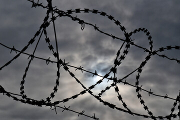 Silhouette of Barbed Wire Against Cloudy Sky with Sun. Barbed Wire Against Overcast Sky with Sun Breaking Through Clouds