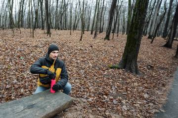 man drinking relaxes, individual resting on wooden seat, person in bright attire pauses for water in city park, urban male in vibrant jacket takes break with drink on leafy ground