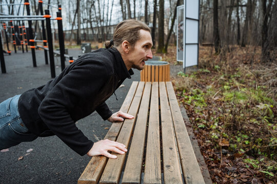 man performing incline pushups outdoors, athletic individual doing incline pushups at park, person executing inclined pushups on wooden bench at outdoor fitness zone in cool weather