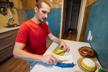 man preparing sandwich with knife and foil on countertop, red shirt and steady hands, fresh loaf and butter visible, compact kitchen setting with livedin textures and everyday comfort