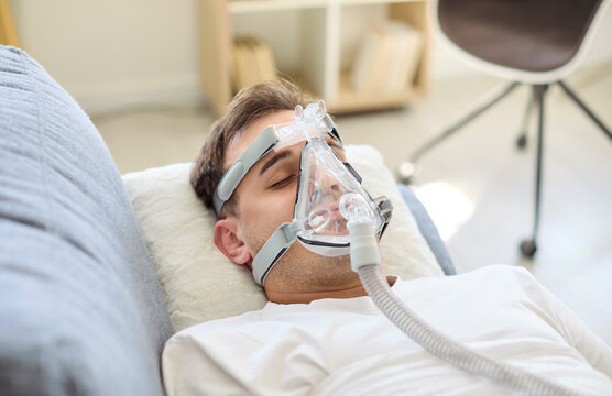 Man in aspirator with oxygen mask resting in bed. The patient gets breathing support during therapy for recovery. Clean setup suggests hospital healthcare at home. Calm care concept of recovery.