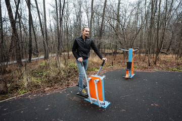 white coach using outdoor elliptical trainer in park, upbeat smile, gray sky and damp ground, demonstration of lowimpact cardio, motivating posture and casual athletic wear