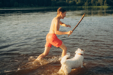 Boy joyfully playing with a dog in the water on a sunny day, holding a stick while the excited dog jumps around. The scene captures the essence of a carefree summer adventure.