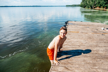 Young person in orange shorts climbs onto a wooden dock from a calm lake. The image captures the moment of climbing, with the serene water and sunny day adding to the summery feeling.