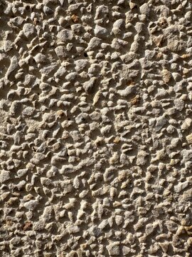 A high-contrast, full-frame macro photograph of a rough-cast mineral wall. The surface is densely packed with small, sharp-edged stones and pebbles embedded in a sandy cement base. The side-angle sunl