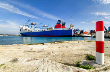 Container ship docked at Birzebbuga port, Malta, with cranes, clear sky, calm sea, and loading zone in background.