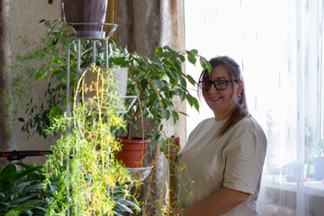 A woman wearing glasses, smiling brightly, is busy tending to her lush collection of houseplants on a stand by the window. The woman demonstrates her passion for gardening by tending to the plants.