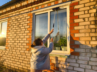 A woman washes a dusty brick window with a long brush during the golden hour, the sun's rays reflecting off the glass. She works outdoors wearing a casual jacket and long sleeves.