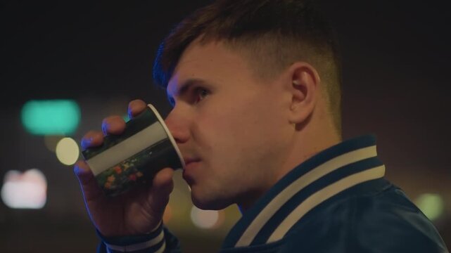 night city profile of man drinking framed by bokeh lights, varsity jacket collar, disposable cup in hand, introspective gaze toward blurred street, cinematic shallow depth, quiet contemplative