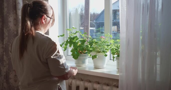 A diligent woman wearing glasses removes potted plants from a windowsill in a sunlit room before beginning spring cleaning and housework. The woman prepares the window for washing.