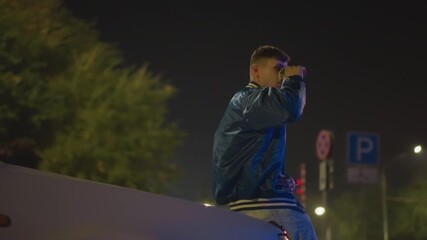 Young white man on car hood at night under neon billboard, wearing glossy bomber jacket and jeans, sipping from takeaway cup, watching empty street, cinematic city lights and quiet parking lot