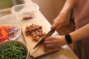 Woman slicing marinated meat on a wooden cutting board next to bowls of chopped red pepper, red onion, and green beans, preparing ingredients for a hearty hot dish